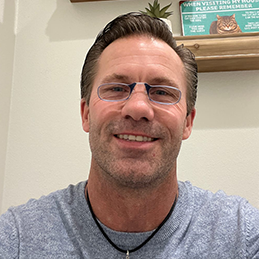 Smiling man with short brown hair and glasses wears a gray shirt. 
